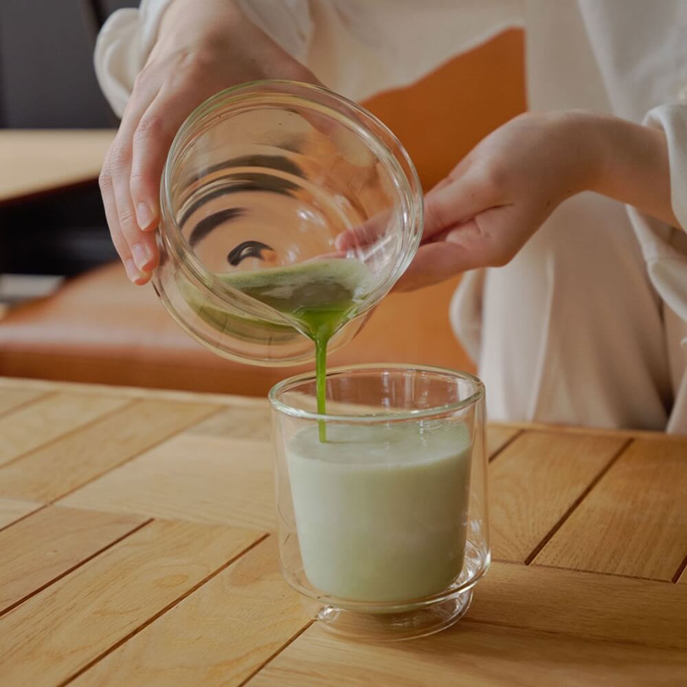 Frisch aufgeschlagener Matcha wird aus einer Glas-Matchaschale mit Ausguss in ein Glas mit Milch gegossen – Matcha Latte Zubereitung